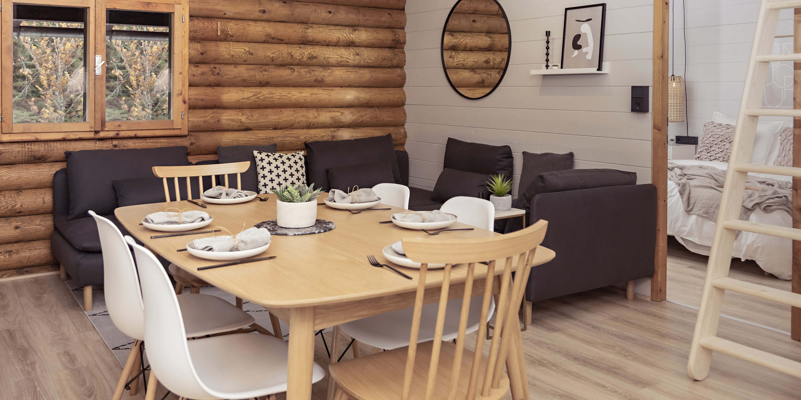 Gedeckter Esstisch für sechs Personen vor der Blockwand im Wohnzimmer von Apartment Frieda, dahinter die Sofalandschaft und die Leiter zum Dachnest.